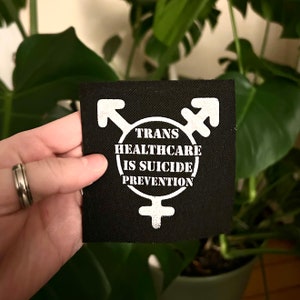 May include: Black fabric patch with white text and symbols. The text reads "Trans Healthcare is Suicide Prevention". The symbols are a transgender symbol and a female symbol.