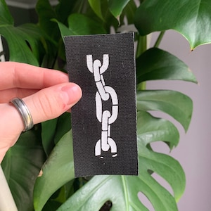 May include: A black fabric patch with a white chain design. The chain is made up of three links.