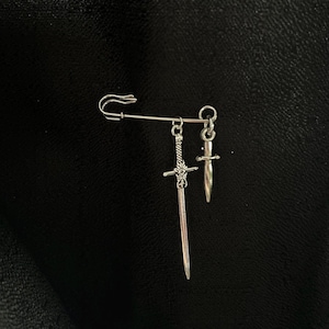 May include: A silver safety pin with two silver sword charms hanging from it. The larger sword charm has a cross guard and a pointed tip. The smaller sword charm is a dagger with a pointed tip.