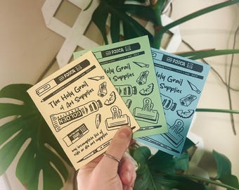 May include: Three art supply guides in yellow, green, and blue are held in a hand. The yellow guide reads "The Holy Grail of Art Supplies" with illustrations of art tools. The green and blue guides say "Holy Grail Supplies".