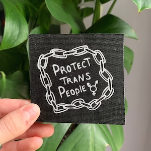 May include: Black fabric patch with white text and a chain border. The text reads "Protect Trans People" with a transgender symbol.