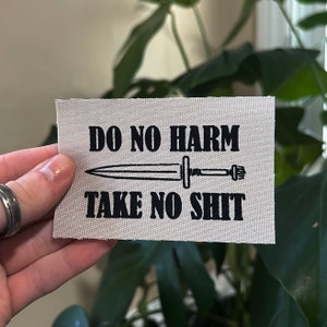 May include: A white fabric patch with black text and a black sword graphic. The text reads "Do No Harm Take No Shit".