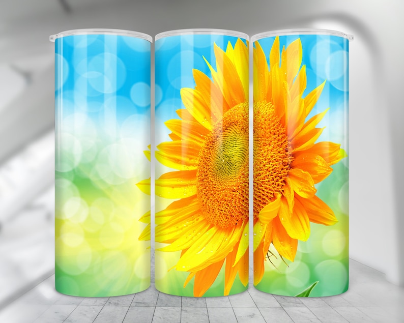 May include: A tumbler with a design of a bright yellow sunflower with a green background and a blue sky with white bokeh circles.