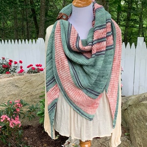 Peut inclure: Un châle triangulaire à rayures dans les tons de vert, rose et gris. Le châle est drapé sur un mannequin blanc.