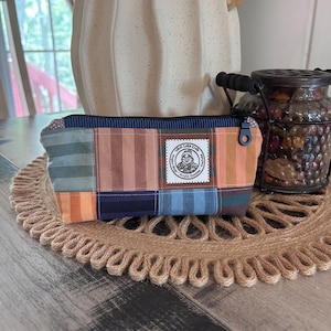 May include: A rectangular fabric zipper pouch featuring a patchwork design of various striped and solid colored panels in shades of blue, orange, brown, and tan. A white square label with black text and a circular illustration is centered on the front.
