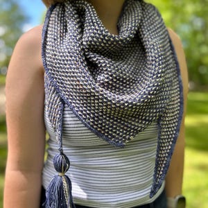 May include: A navy and cream knitted shawl with a textured pattern. The triangular shawl has a navy border and a tassel of navy and cream yarn. The person is wearing a striped vest top and blue jeans. The shawl's dimensions are approximately 150 cm x 75 cm.