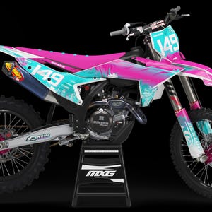Puede incluir: Una moto de motocross rosa y turquesa con el número 149 en el lateral. La moto está sobre un soporte negro y tiene un fondo negro.