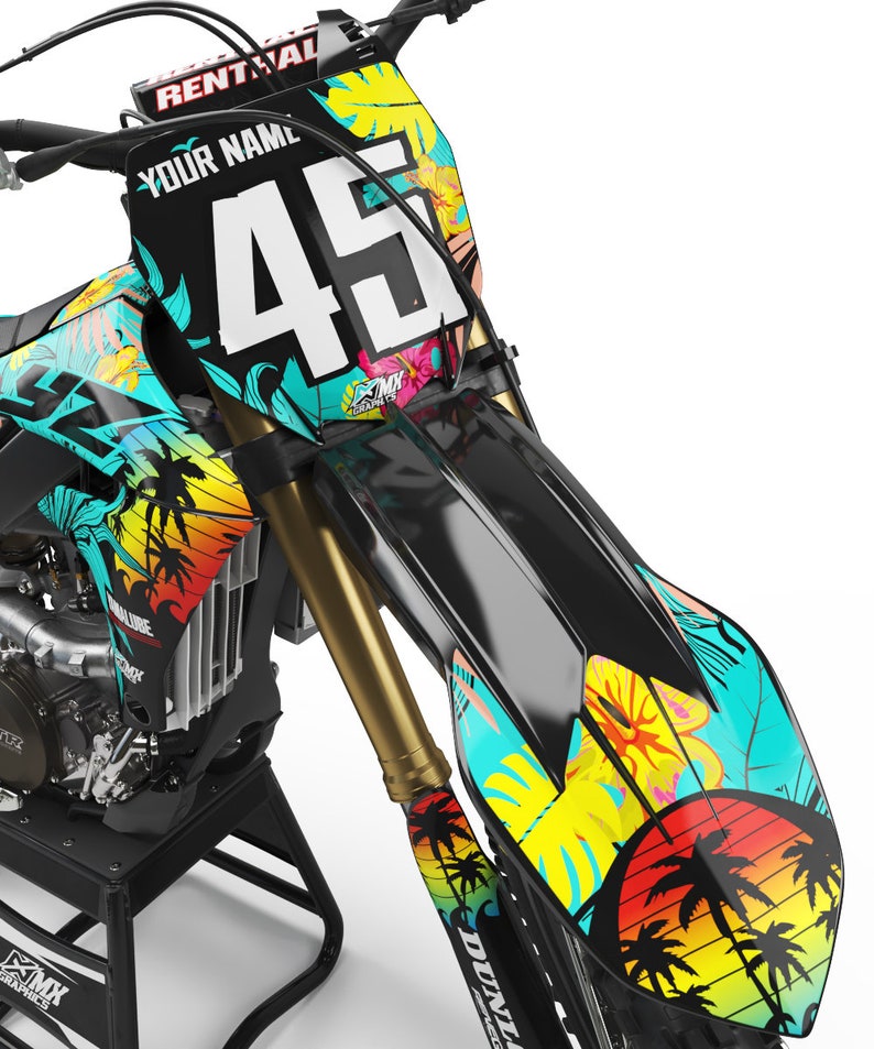 May include: A black and turquoise motocross bike with a tropical print design. The bike has a number plate with the number 45 and the words "Your Name" on it. The bike is on a stand and has a yellow and orange sunset graphic on the front fender.