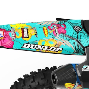 May include: A turquoise and black dirt bike with floral graphics. The graphics feature pink hibiscus flowers, yellow and white leaves, and the words "Dunlop", "VMXALUBE", "FREAKSPIX", "100%", and "RENEGADE".