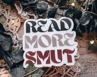 May include: A white sticker with the words "READ MORE SMUT" in a retro font. The text is layered in black, gray, and burgundy. The sticker is surrounded by greenery, a crocheted doily, and fairy lights.