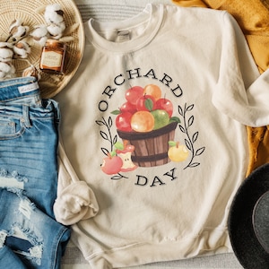 Pode incluir: Uma camisa de moletom bege com o texto "ORCHARD DAY" e uma ilustra&ccedil;&atilde;o em aquarela de uma cesta de ma&ccedil;&atilde;s e laranjas.