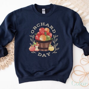 Pode incluir: Camisola azul marinho com um gr&aacute;fico de uma cesta de ma&ccedil;&atilde;s e o texto "Orchard Day".