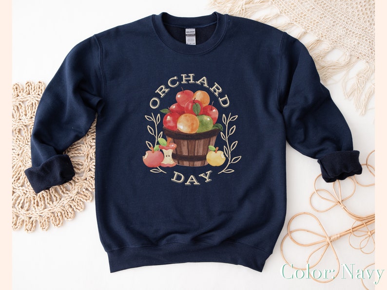 Pode incluir: Camisola azul marinho com um gr&aacute;fico de uma cesta de ma&ccedil;&atilde;s e o texto "Orchard Day".