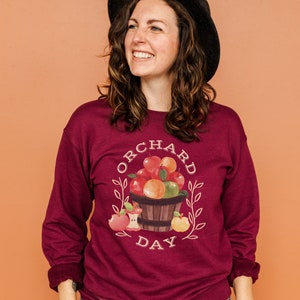 Pode incluir: Uma camisa de manga comprida de cor vinho com um gr&aacute;fico branco de uma cesta de ma&ccedil;&atilde;s e o texto "ORCHARD DAY".