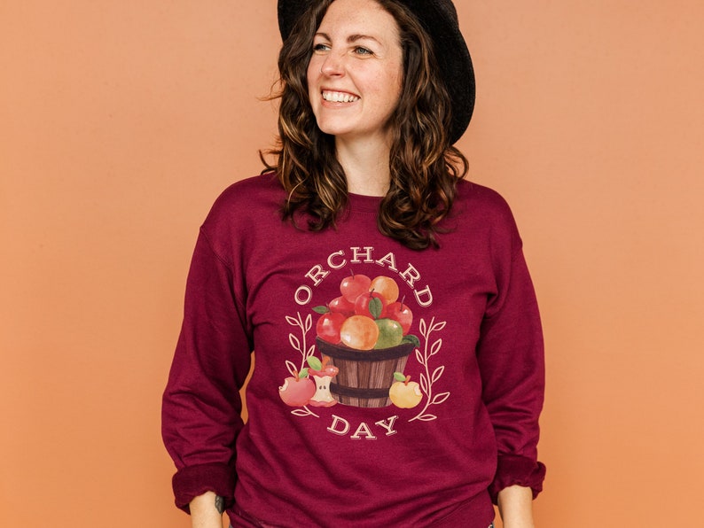 Pode incluir: Uma camisa de manga comprida de cor vinho com um gr&aacute;fico branco de uma cesta de ma&ccedil;&atilde;s e o texto "ORCHARD DAY".