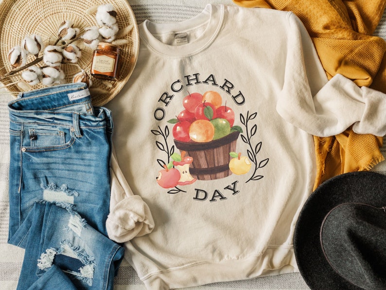 Pode incluir: Uma camisa de moletom bege com o texto "ORCHARD DAY" e uma ilustra&ccedil;&atilde;o em aquarela de uma cesta de ma&ccedil;&atilde;s e laranjas.