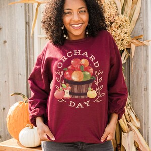 Pode incluir: Uma camisa de moletom vinho com um gr&aacute;fico de uma cesta de ma&ccedil;&atilde;s e o texto "Orchard Day".