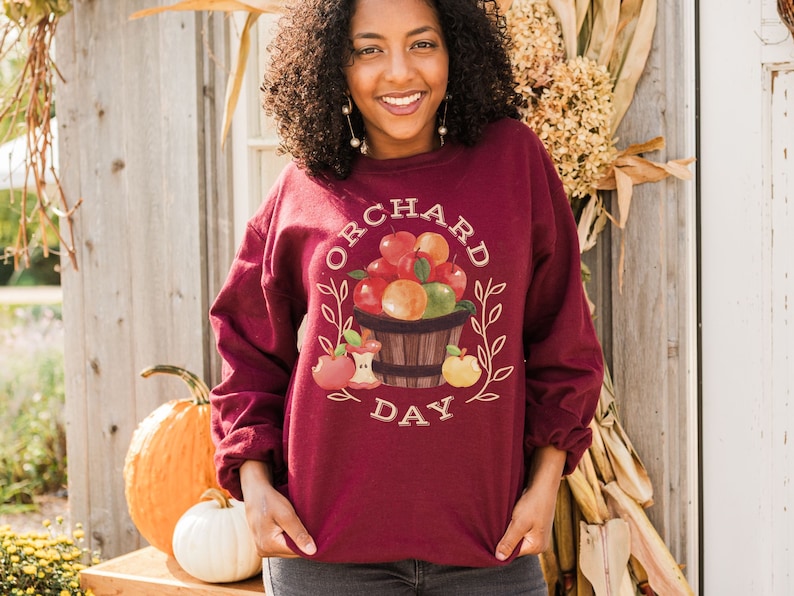 Pode incluir: Uma camisa de moletom vinho com um gr&aacute;fico de uma cesta de ma&ccedil;&atilde;s e o texto "Orchard Day".