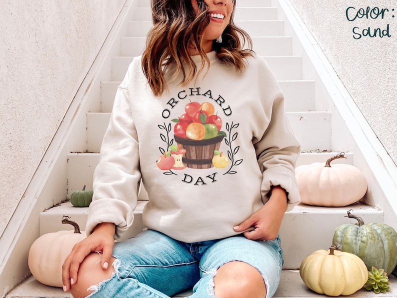 Pode incluir: Uma camisa de moletom cor areia com um gr&aacute;fico de uma cesta de ma&ccedil;&atilde;s e o texto "ORCHARD DAY".
