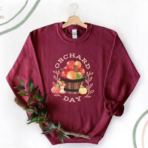 Pode incluir: Uma camisa de moletom vinho com um gr&aacute;fico branco de uma cesta de ma&ccedil;&atilde;s e o texto "Orchard Day".