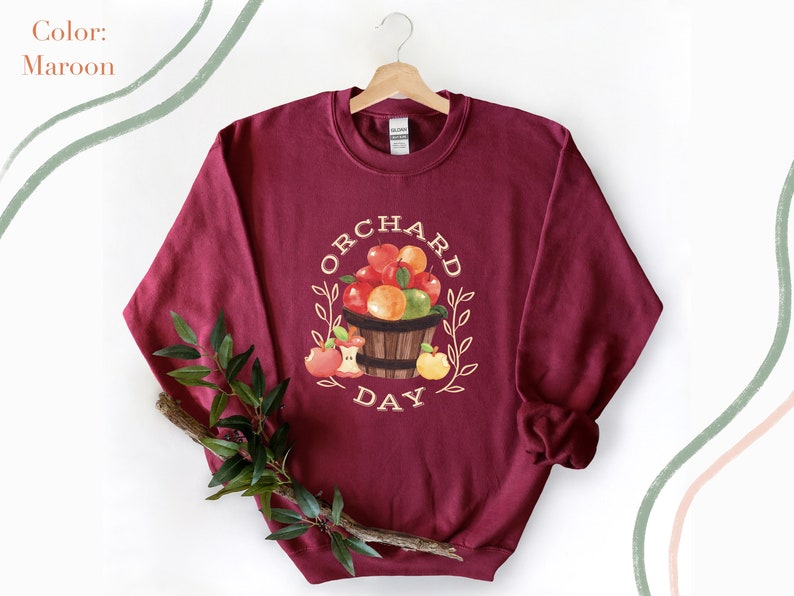 Pode incluir: Uma camisa de moletom vinho com um gr&aacute;fico branco de uma cesta de ma&ccedil;&atilde;s e o texto "Orchard Day".