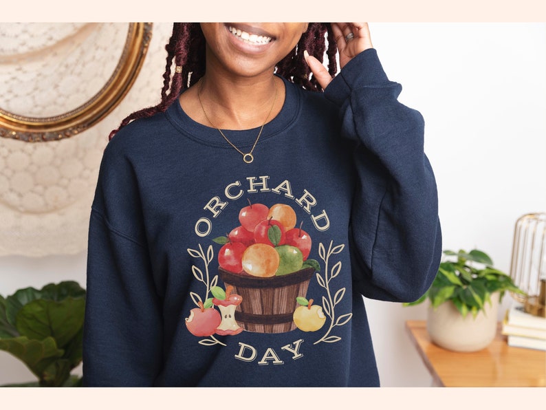 Pode incluir: Uma camisa de moletom azul marinho com um gr&aacute;fico de uma cesta de ma&ccedil;&atilde;s e o texto "ORCHARD DAY" em uma fonte de estilo vintage.
