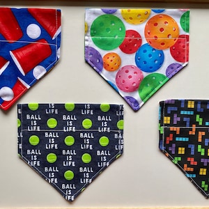 May include: Four different dog bandanas with various patterns. The first bandana is blue with red solo cups and white polka dots. The second bandana is white with multi-colored pickleball designs. The third bandana is dark blue with green tennis balls and the text "Ball is Life". The fourth bandana is black with a pattern of colorful Tetris blocks.