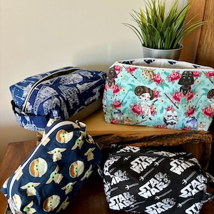 Puede incluir: Cuatro bolsas de cremallera de tela diferentes. Una bolsa es negra con un estampado blanco de "Star Wars". Otra bolsa es negra con un estampado verde de "Baby Yoda". Una tercera bolsa es azul con un estampado blanco de "Star Wars". La última bolsa es azul claro con un estampado floral y una imagen de dibujos animados de la princesa Leia.