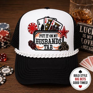 May include: A black and white trucker hat with a white front panel featuring playing cards, dice, and the text "PUT IT ON MY HUSBANDS TAB." A red star and poker chips are also displayed. The hat has a white rope trim and a black mesh back.