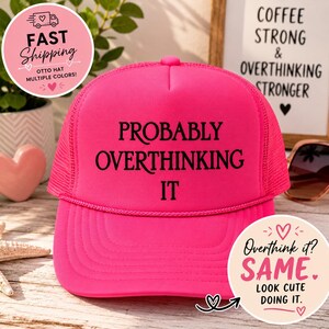 May include: A bright neon pink trucker hat featuring a mesh back, a foam front panel, and a decorative cord across the brim. The text PROBABLY OVERTHINKING IT is printed in black, bold, serif capital letters across the center of the front panel.