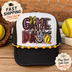 Op de afbeelding: Een zwart-witte truckerpet met een grafische print met de tekst GAME DAY in dikke letters met luipaard-, zebra- en ruitpatronen. De pet is versierd met een rij kleine gele softball-kralen langs de voorzijde van de klep.