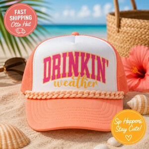 Peut inclure: Une casquette de type trucker couleur pêche avec un panneau avant blanc portant l'inscription DRINKIN' weather en lettres roses et jaunes. La casquette présente une chaîne décorative en plastique couleur pêche à la base du panneau avant.