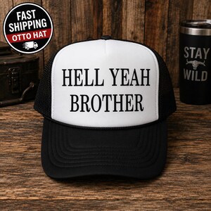 Pode incluir: Um boné trucker preto e branco com um painel frontal branco apresentando o texto preto HELL YEAH BROTHER impresso em uma fonte serifada em negrito. O boné inclui uma aba preta, uma parte traseira de malha preta e um detalhe de cordão preto.