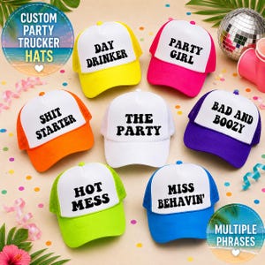 May include: A selection of vibrant trucker hats with white fronts and mesh backs. Each hat displays a different phrase in bold black text, such as "Day Drinker," "Party Girl," and "Hot Mess." The hats are arranged on a light surface with party decorations.
