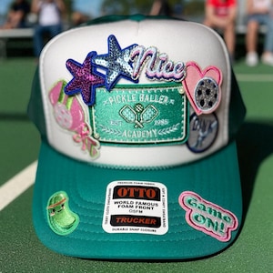 May include: A teal and white trucker hat with multiple embroidered patches. Patches include stars, hearts, and the words "Nice" and "Game On!". The hat features the text "Pickle Baller Academy" and "1985".