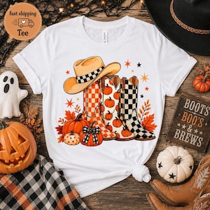 May include: A white short-sleeve t-shirt featuring a graphic of a tan cowboy hat and three western boots. The boots display orange checkered, pumpkin, and black checkered patterns. Orange pumpkins, autumn leaves, and stars surround the boots.