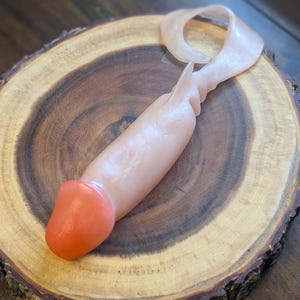 May include: A flesh-toned sex toy with an orange tip and a loop at the top. The toy is lying on a round, wooden surface with a dark brown center and a lighter brown edge.