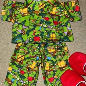 May include: Green pajama set with a repeating pattern of Teenage Mutant Ninja Turtles characters. The set includes a short-sleeved top and matching shorts. Two red slippers are next to the pajama set.