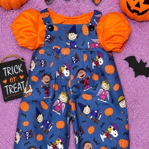 May include: A blue overall-style outfit with a Peanuts Halloween print featuring characters in costumes, orange pumpkins, and bats. The garment has short orange puff sleeves, two black buttons at the shoulders, and includes two matching hair bows.