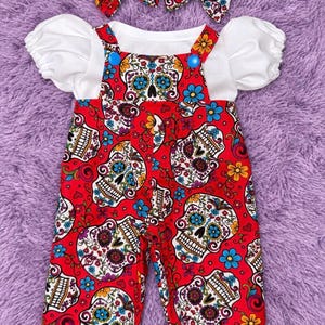 May include: A red overall-style outfit featuring a colorful sugar skull pattern with white, blue, yellow, and purple accents. The garment includes a white short-sleeved shirt with puff sleeves, two bright blue buttons, and two matching hair bows.