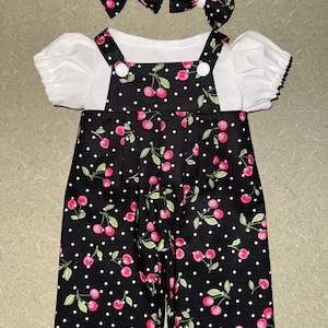 May include: A baby outfit set featuring a white short-sleeved top and black overalls. The overalls are adorned with a cherry and polka dot pattern. Two matching bow hair accessories complete the ensemble.