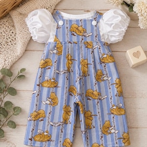 May include: A blue and white striped overall dress featuring a repeating pattern of a yellow bear character. The garment includes a white short-sleeved top with puffed sleeves attached underneath, secured with white buttons at the shoulder straps.
