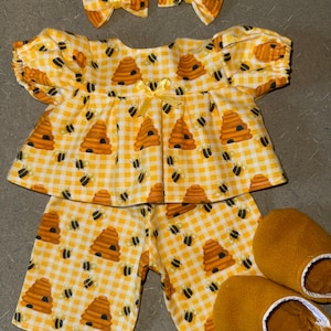 May include: A yellow and white gingham pajama set with a bee and beehive pattern. The set includes a top, shorts, two hair bows, and a pair of matching slippers. The top has short sleeves and a yellow bow.