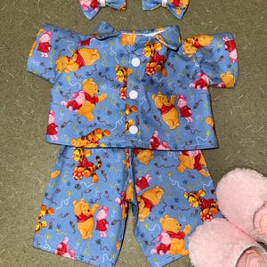 May include: A light blue pajama set featuring Winnie the Pooh, Piglet, and Tigger characters. The set includes a button-down shirt, shorts, and two matching bow hair clips. Pink fuzzy slippers are also included.