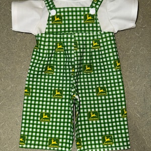 May include: A two-piece outfit featuring a white t-shirt and green and white checkered overalls. The overalls have the John Deere logo printed throughout. The outfit is designed for a baby or toddler.
