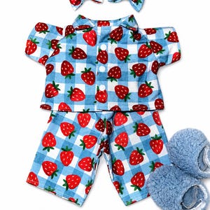May include: A blue and white gingham pajama set with red strawberries, including a short-sleeved top, shorts, two matching bow hair clips, and blue fuzzy slippers. The pajamas have a button-down front.
