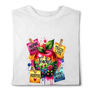 May include: White t-shirt featuring a colorful apple design with the word "Teacher" in the center. The apple is surrounded by flowers and leaves, with the words "Inspire Minds", "Teach Love", "Mentor", and "Difference Maker" on attached notes.