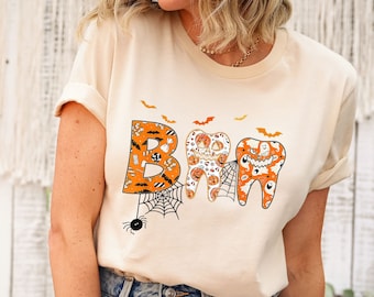 May include: A cream colored t-shirt with a graphic of three teeth decorated with Halloween themed designs. The teeth spell out the word "BOO" with bats flying above them.