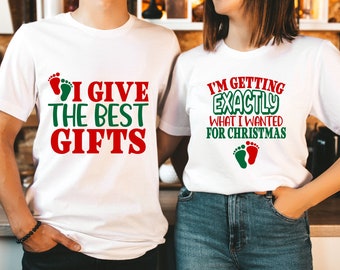 May include: Two white t-shirts with a Christmas theme. The first shirt says "I give the best gifts" with a green and red footprint. The second shirt says "I'm getting exactly what I wanted for Christmas" with a green and red footprint.