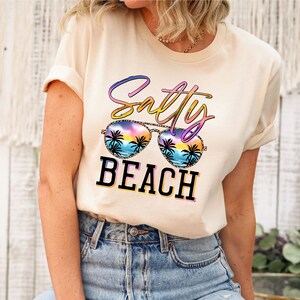 May include: A cream-colored t-shirt with the words "Salty Beach" in a colorful design. The sunglasses graphic features a beach scene with palm trees and a sunset. The shirt is paired with blue jeans.
